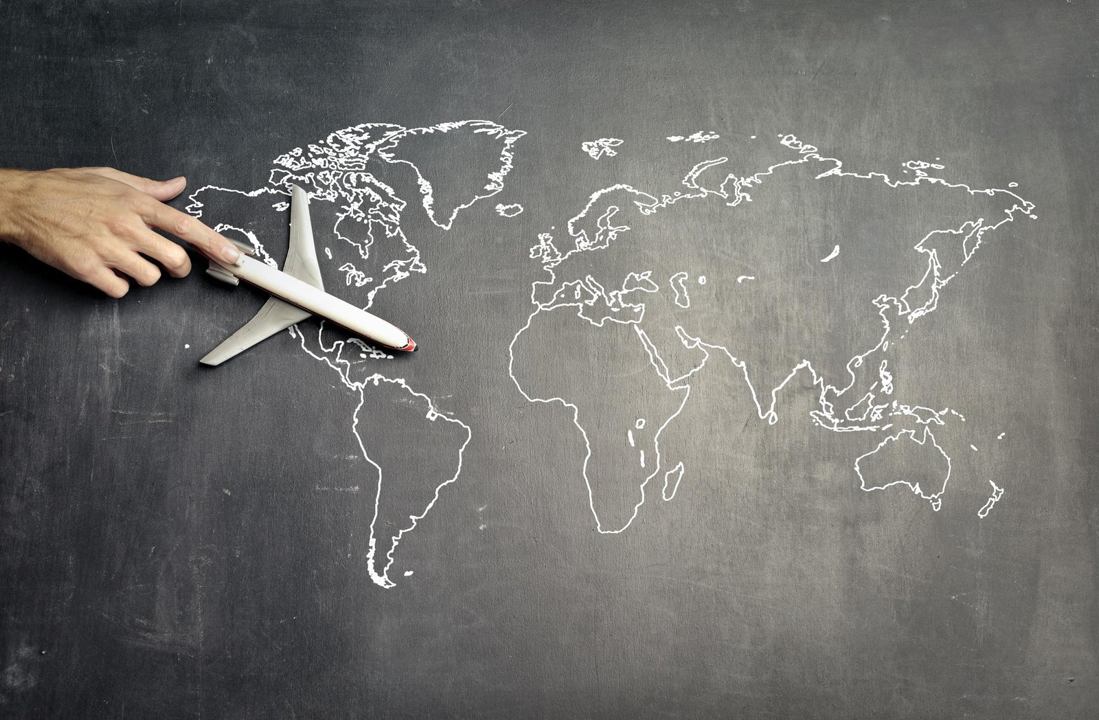 From above of crop anonymous person driving toy airplane on empty world map drawn on blackboard representing travel concept