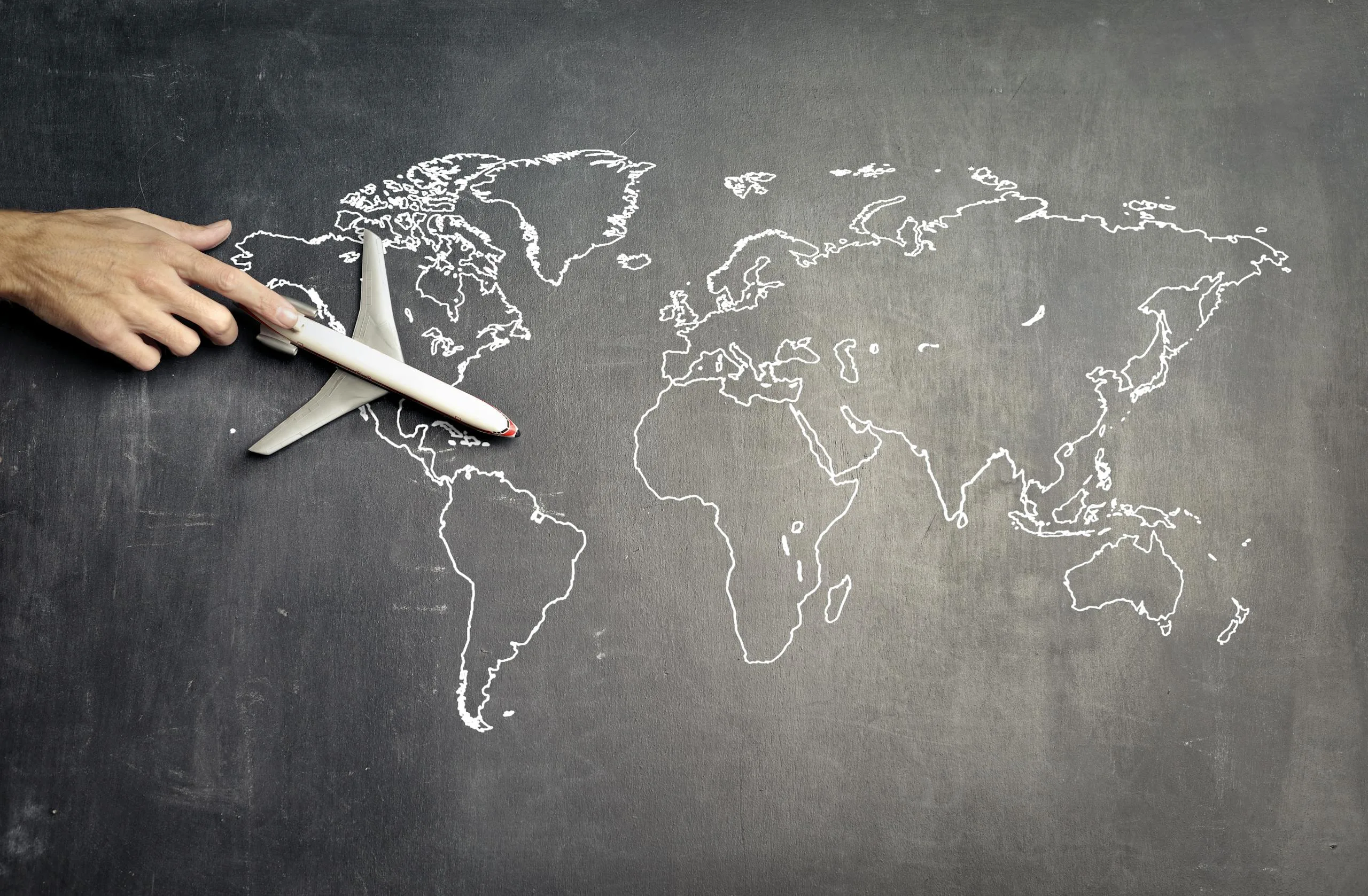 From above of crop anonymous person driving toy airplane on empty world map drawn on blackboard representing travel concept