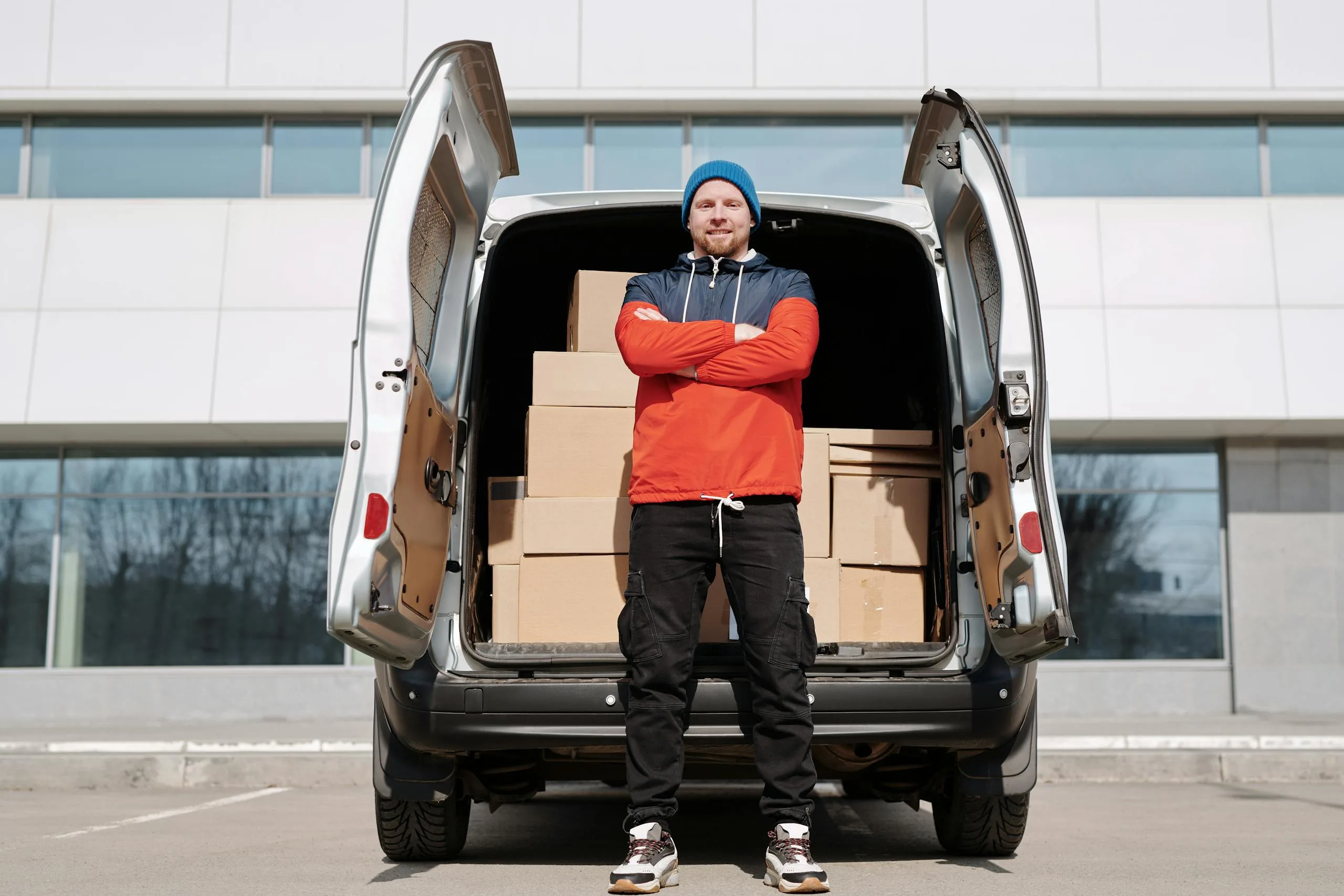 Man in red jacket and blue beanie stands by van of boxes in the city, ready for delivery.
